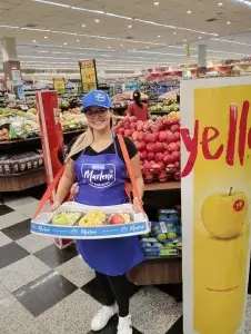 SP< Uniformes - Mulher em supermercado vestindo boné e avental promocional de empresa, sobre camiseta gola V e calça, segurando bandeja de degustação de divulgação de produtos.
