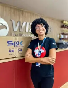 SP< Uniformes - Foto de mulher cliente vestindo uniforme da empresa Wizard ao lado de caixa com itens personalizados entregues pela SP< Uniformes.